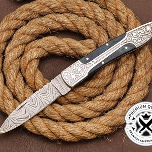May include: A silver pocket knife with a patterned blade and a dark blue handle with silver accents. The knife is lying on a brown rope. The text "PREMIUM QUALITY" is on a black circle with a crown and crossed swords. The text "ATLANTA CUSTOM BLADES" is below the circle.