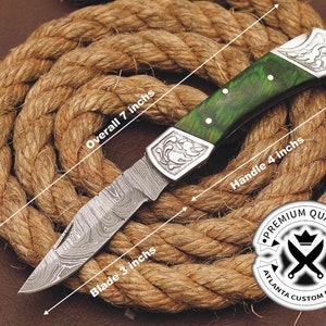May include: A green and silver pocket knife with a Damascus steel blade, a green wood handle, and a silver floral design on the handle. The knife is 7 inches long, the blade is 3 inches long, and the handle is 4 inches long. The knife is on a brown rope background. The text "PREMIUM QUALITY ATLANTA CUSTOM BLADES" is on a white circle with a black crown and crossed swords.