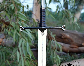 May include: A silver sword with a black handle and a gold pommel. The blade is inscribed with the text "↑日 K1141 Bnri ΝΗΜΙΚ *INKY&BN". The sword is in front of a green leafy background.