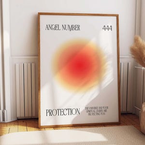 May include: A white poster with a brown wooden frame. The poster has a red and orange gradient circle in the center. The text "ANGEL NUMBER 444" is at the top of the poster. The text "PROTECTION" is at the bottom left of the poster. The text "THE UNIVERSE AND YOUR SPIRITUAL GUIDES ARE PROTECTING YOU" is at the bottom right of the poster.