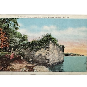 May include: Vintage postcard featuring the Ruins of Fort Frederica on Saint Simons Island, GA. The image shows a stone structure covered in greenery, situated near a body of water. The sky is a mix of blue and orange hues, and trees are visible on the left.