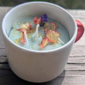 May include: A light blue candle poured into a white ceramic mug with a red handle. The candle surface is decorated with scattered dried flower petals in shades of pink, yellow, and purple, along with small yellow beads surrounding a central wick.