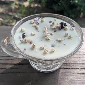 May include: A pale green candle in a clear glass teacup with a handle. The wax surface is embedded with small dried white flowers, dark blue flower petals, and tiny purple crystal chips, centered around a single white cotton candle wick.