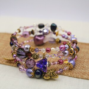 May include: A multi-strand wrap bracelet with purple, lavender, and clear beads. The bracelet features round, faceted, and floral beads, and a gold-coloured butterfly charm. The bracelet is approximately 17.8 cm in circumference.