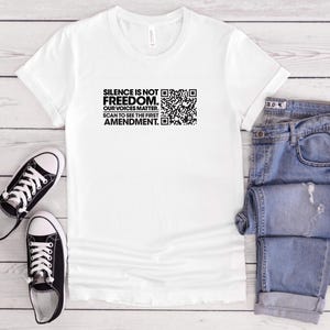 Può includere: Una t-shirt bianca con testo nero che recita SILENCE IS NOT FREEDOM. OUR VOICES MATTER. SCAN TO SEE THE FIRST AMENDMENT. accanto a un codice QR in bianco e nero. La maglietta è mostrata insieme a un paio di scarpe da ginnastica nere e jeans blu.