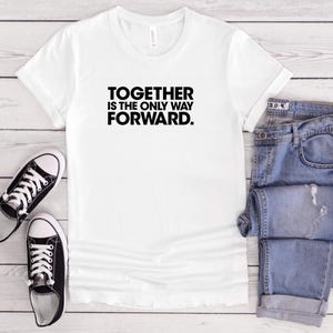 May include: White t-shirt with the text "TOGETHER IS THE ONLY WAY FORWARD" in black. The shirt is paired with blue jeans and black and white sneakers. The items are arranged on a light wood surface.