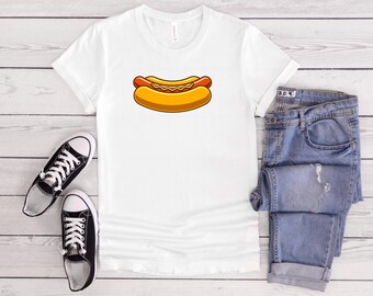 May include: White t-shirt featuring a cartoon hot dog design. The hot dog has a red sausage, yellow mustard, and a yellow bun. The image also shows a pair of black sneakers and blue jeans.