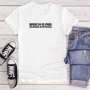 May include: A white t-shirt with the black text "SPEECH IS FREE. LISTENING IS OPTIONAL." is displayed with blue jeans and black and white trainers. The items are arranged on a light wood-grain surface.