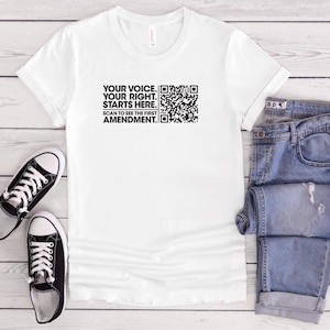 May include: White t-shirt with the text "YOUR VOICE. YOUR RIGHT. STARTS HERE. SCAN TO SEE THE FIRST AMENDMENT." and a QR code. The shirt is paired with blue jeans and black and white sneakers.