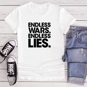 May include: White t-shirt with the text "ENDLESS WARS. ENDLESS LIES." in bold black letters. The shirt is paired with blue jeans and black and white sneakers.