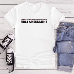 May include: White t-shirt with the text "SPEAK HONESTLY OPENLY UPHOLDING TRUTH FIRST AMENDMENT" in black and red. The shirt is paired with blue jeans and black and white trainers on a light wood surface.