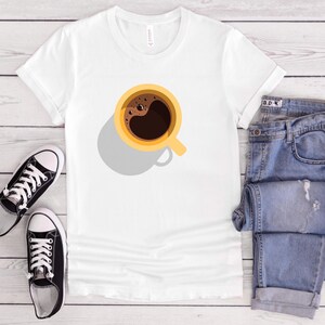Puede incluir: Camiseta blanca con un diseño de taza de café, combinada con vaqueros azules y zapatillas negras. La taza de café es amarilla con un diseño de café marrón oscuro. Los vaqueros están doblados y las zapatillas son negras y blancas.