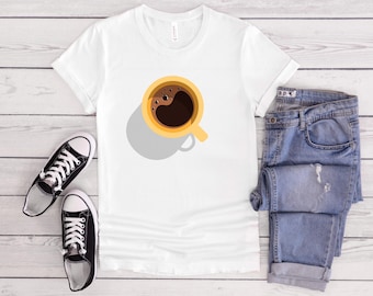 Puede incluir: Camiseta blanca con un diseño de taza de café, combinada con vaqueros azules y zapatillas negras. La taza de café es amarilla con un diseño de café marrón oscuro. Los vaqueros están doblados y las zapatillas son negras y blancas.
