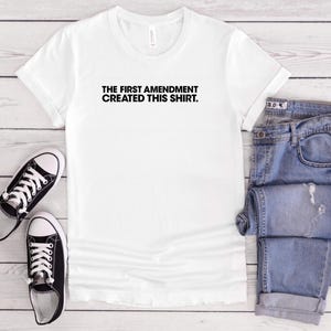 Könnte beinhalten: Weißes T-Shirt mit dem Text "THE FIRST AMENDMENT CREATED THIS SHIRT." Das Bild zeigt auch schwarz-weiße Turnschuhe und eine blaue Jeans. Das T-Shirt ist aus einem weichen Material.