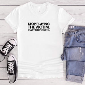 May include: A white short-sleeved t-shirt featuring the black text "STOP PLAYING THE VICTIM. START GOVERNING." printed on the chest, alongside a pair of blue denim jeans and two black and white low-top canvas trainers.