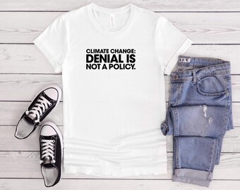 May include: White t-shirt with the text "CLIMATE CHANGE: DENIAL IS NOT A POLICY" in black. The shirt is paired with blue jeans and black and white sneakers. The items are arranged on a light wood-grain surface.