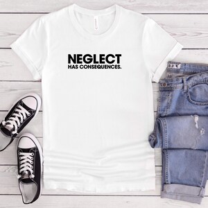 Puede incluir: Una camiseta blanca con la frase "NEGLECT HAS CONSEQUENCES" en negro. La imagen también muestra unos vaqueros azules y zapatillas deportivas en blanco y negro. Los artículos están colocados sobre una superficie clara con vetas de madera.