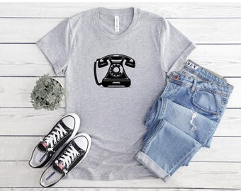 May include: A light gray t-shirt featuring a black graphic of a vintage rotary phone. The shirt is paired with light blue denim jeans and black and white sneakers. A small potted plant is also visible.