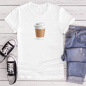 Peut inclure: Un t-shirt blanc avec un motif de tasse à café, associé à un jean bleu et des baskets noires. La tasse à café est marron et blanche. Le jean est plié. Les baskets sont noires et blanches.