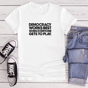 May include: A white short-sleeved t-shirt featuring the black text "DEMOCRACY WORKS BEST WHEN EVERYONE GETS TO PLAY." accompanied by a pair of black and white low-top canvas sneakers and a pair of blue denim jeans with distressed detailing.