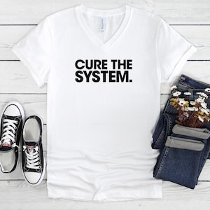 以下が含まれることがあります： 「CURE THE SYSTEM」の黒い文字がプリントされた白いVネックTシャツ。 黒と白のスニーカーと、花が飾られた青いジーンズが写っています。
