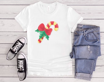 May include: White t-shirt with a red and green candy cane with a red bow on top. The candy cane is on a white background.