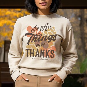 May include: A woman wearing a beige sweatshirt with the text "In All Things Give Thanks 1 Thessalonians 5:18" in brown lettering with a fall leaf design. She is standing in front of a wall of pumpkins.