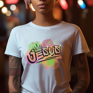 May include: A white t-shirt with a colorful graphic design of the word "JESUS" with a crown above it. The graphic is surrounded by pink, yellow, and green watercolor-like splatters.