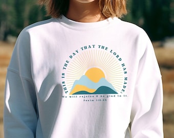 May include: White sweatshirt with a graphic design of a mountain range with a sun and the text "THIS IS THE DAY THAT THE LORD HAS MADE, We will rejoice & be glad in it. Psalm 118:24"