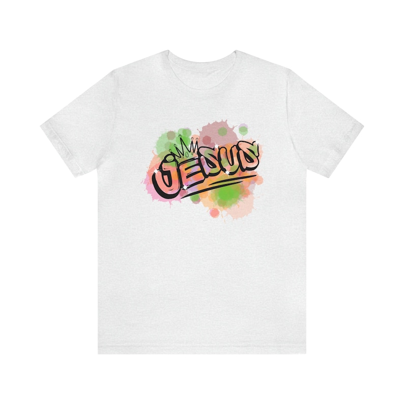 May include: White t-shirt with a colorful watercolor design of the word "JESUS" with a crown above it.