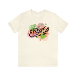 May include: A white t-shirt with a colorful graphic of the word "JESUS" with a crown above it. The graphic is surrounded by watercolor-like splatters of green, pink, and orange.