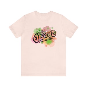 May include: A light pink t-shirt with a colorful graphic of the word "JESUS" with a crown above it. The graphic is surrounded by watercolor-like splatters of pink, green, and orange.