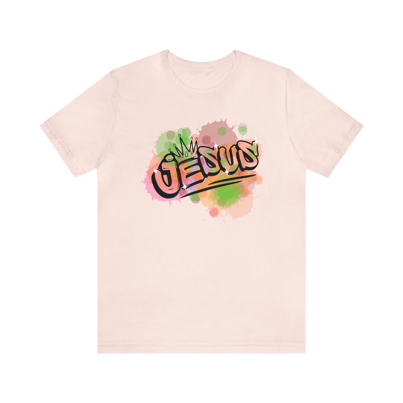 May include: A light pink t-shirt with a colorful graphic of the word "JESUS" with a crown above it. The graphic is surrounded by watercolor-like splatters of pink, green, and orange.