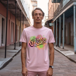 May include: A pink t-shirt with a graphic design of the word "JESUS" in a graffiti style with a crown above the word. The graphic is surrounded by colorful watercolor-like splatters.