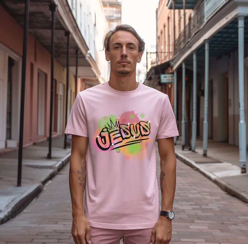 May include: A pink t-shirt with a graphic design of the word "JESUS" in a graffiti style with a crown above the word. The graphic is surrounded by colorful watercolor-like splatters.