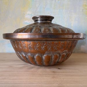 May include: A copper-colored bowl with a matching lid. The bowl has a rounded shape with a decorative floral pattern around the base. The lid has a knob on top and a ridged design. The bowl is sitting on a wooden surface.