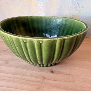 May include: A green ceramic bowl with a ribbed exterior. The bowl has a glossy finish and a slightly irregular rim. The bowl is sitting on a wooden surface.