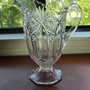 May include: Clear glass pitcher with a decorative starburst and geometric pattern. The pitcher has a handle and a spout, and sits on a pedestal base. The pitcher is likely used for serving beverages.