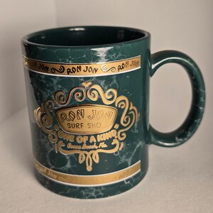 May include: A dark green ceramic mug with a marble effect. The mug features gold lettering and accents, including the text "RON JON SURF SHOP" and "ONE OF A KIND, Cocoa Beach, FL". The handle is also dark green.