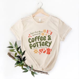 May include: A beige t-shirt with a green and orange graphic that says "Today's Good Mood Sponsored By Coffee & Pottery" with a coffee cup, coffee beans, and a flower design.