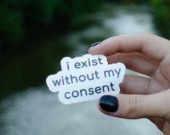 May include: A white sticker with the text "i exist without my consent" held in a hand with black nail polish. The sticker is in focus, while the background is blurred and shows a green forest and a river.