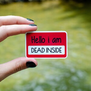 May include: A white sticker with red text that reads "Hello i am DEAD INSIDE" held by a hand with black nail polish.
