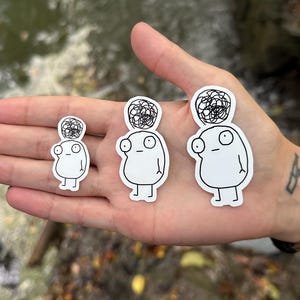 May include: Three white stickers featuring black line drawings of a cartoon character with a messy bun on their head. The character has large eyes and a simple body. The stickers are of varying sizes.