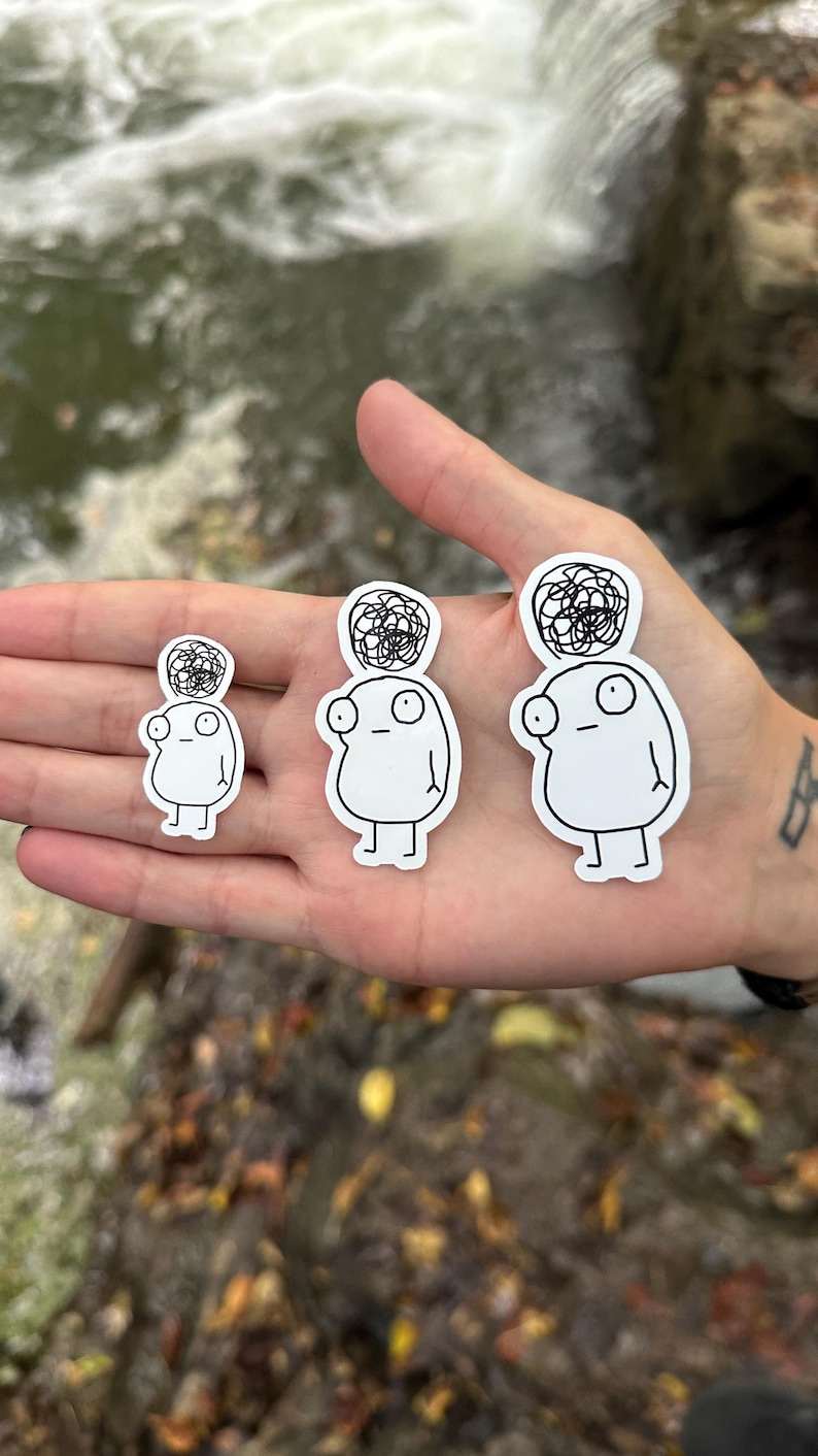 May include: Three white stickers featuring black line drawings of a cartoon character with a messy bun on their head. The character has large eyes and a simple body. The stickers are of varying sizes.