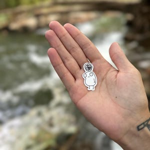 May include: A white sticker of a simple cartoon character with a black and white patterned head. The character has a round body, two small arms, and two small legs.
