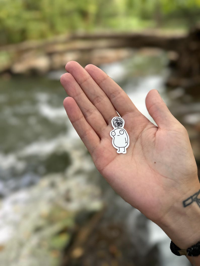 May include: A white sticker of a simple cartoon character with a black and white patterned head. The character has a round body, two small arms, and two small legs.