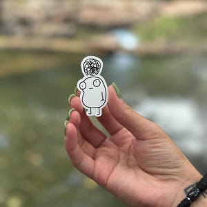 May include: A white sticker with a simple line drawing of a person with a messy bun on their head. The person has large eyes and a small mouth. The sticker is being held by a hand.