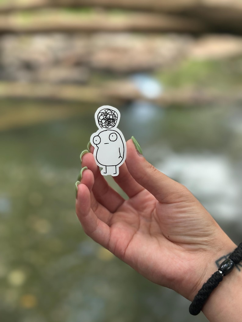 May include: A white sticker featuring a simple line drawing of a person with a messy bun on their head. The person has large eyes and a small mouth. The sticker is being held by a hand.