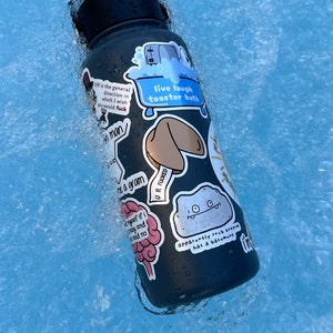 May include: A black water bottle with various stickers on it, including a fortune cookie, a brain, a rock with a basement, and a toaster bath. The bottle is floating in a blue pool of water.