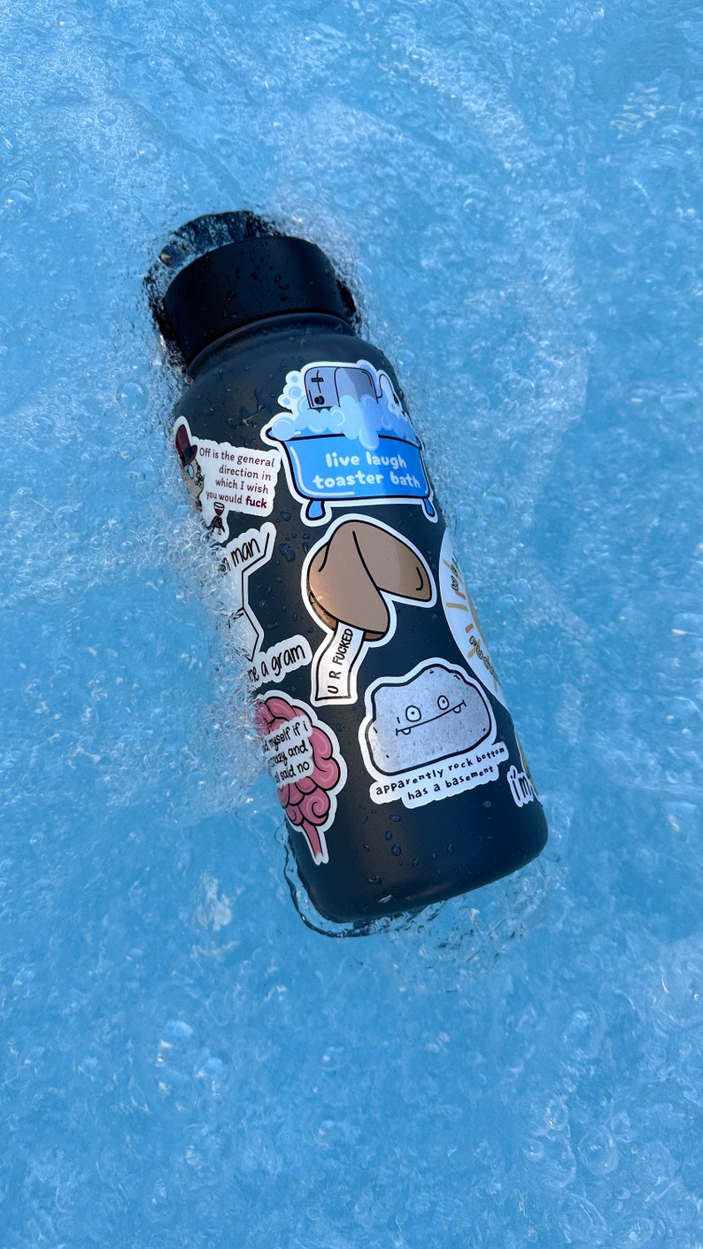 May include: A black water bottle with various stickers on it, including a fortune cookie, a brain, a rock with a basement, and a toaster bath. The bottle is floating in a blue pool of water.